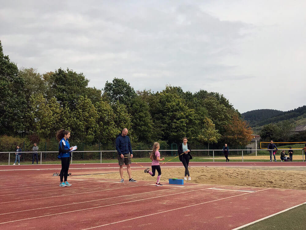 Kinder und betreuende Erwachsene auf der Laufbahn im Stadion mit grünen Hügeln im Hintergrund